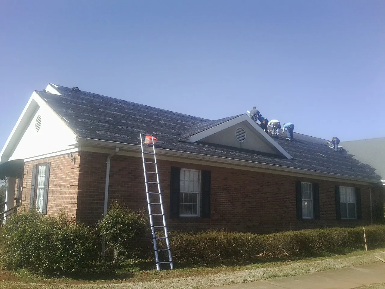 Crew tearing shingles off a brick ranch roof with a ladder set — Metal Roof Repair work by our crew in Port Edwards