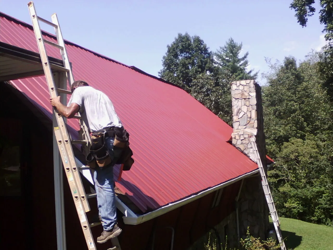Roofer on a ladder beside a red metal roof and stone chimney — Soffit & Fascia Repair by Roofing Brothers in Port Edwards
